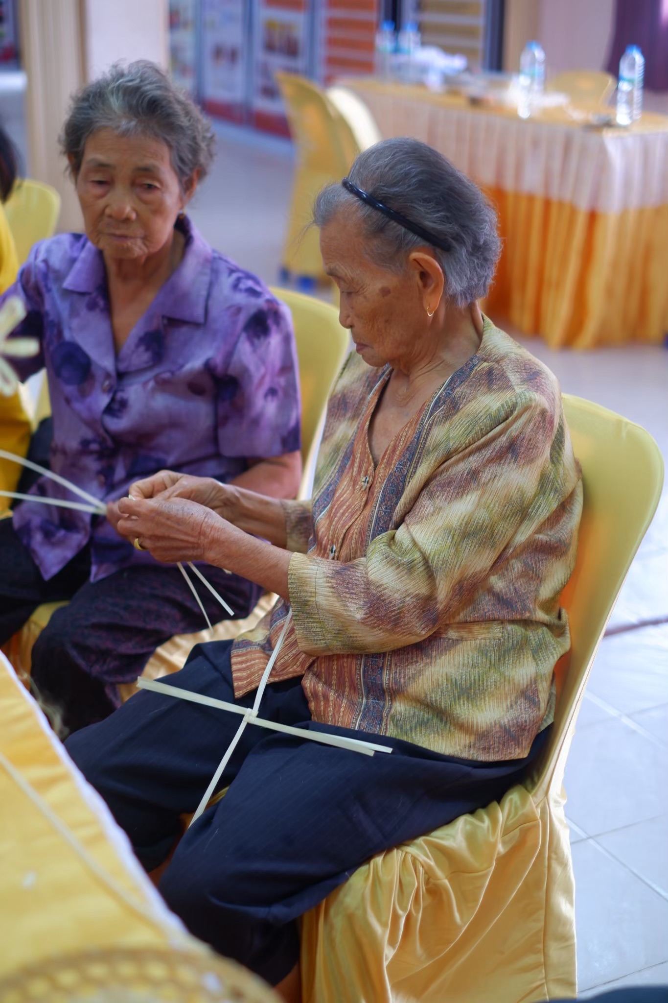 ผู้สูงอายุสองท่านร่วมกิจกรรมจักสาน เห็นใบหน้าและมือของผู้สูงอายุด้านหน้าขณะจับจัดเส้นตอก